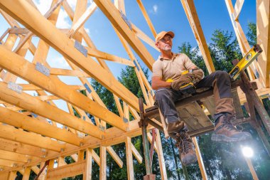 Bir inşaat işçisi, bir yapının ortasında tahta bir kirişin üzerinde otururken mola verir. Gökyüzü açık ve ağaçlar çevrenin çevresini sarıp yeşillik ekliyor..