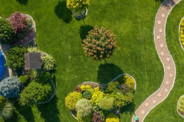 Bahçe, gür yeşillik, renkli çiçekler ve bir ağacın altında gölgeli bir taraçaya uzanan dolambaçlı bir çakıl yolu ile karmaşık bir tasarım içerir..