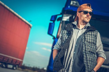 Bir kamyon şoförü mavi kamyonetinin yanında, güneş gözlüğü ve şapka takmış, açık gökyüzüne karşı sıradan bir hava sergiliyor..
