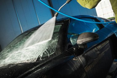 Bir insan siyah bir arabayı yıkamak için yüksek basınçlı bir hortum kullanır, ön cama odaklanır. Su spreyleri iyi aydınlatılmış bir araba yıkamada ferahlatıcı bir sahne yaratır..