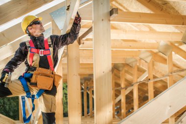 Güvenlik donanımlı bir inşaat işçisi ahşap bir yapıdaki kirişleri ayarlıyor. Parlak güneş ışığı kirişlerin arasından akıyor, odaklanmasını vurguluyor..