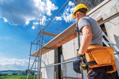 Kask ve alet kemeri takan bir inşaat işçisi inşaat halindeki bir binayı teftiş ederken, güneşli bir günde inşaat iskelesinin çevresi sarıldı..