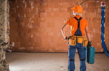 Turuncu kasklı ve gömlekli bir inşaat işçisi boş bir odada duruyor, tadilata hazır. Duvarlar korunmasız malzemelerle dolu..