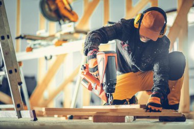 Odaklanmış bir işçi çivi tabancasını ustalıkla kullanarak tahta bir yüzeye diz çöker. İnşaat alanı, doğal ışık tarafından aydınlatılan aletler ve ahşap çerçevelerle dolu..