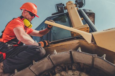 Güvenlik teçhizatlı bir inşaat işçisi açık mavi gökyüzünün altında gün ışığında odaklanmış ve bakımlı bir buldozerin bakımını ve denetimini yapıyor..