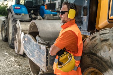 Bir inşaat işçisi, yoğun bir inşaat alanında kısa bir süre dinlenmek için güvenlik teçhizatı ve güneş gözlüğü takarak ağır makinelerin yanında durur..