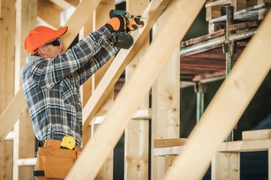 Turuncu şapkalı ve güneş gözlüklü bir işçi şantiyede ahşap kirişleri deliyor. Yoğun ortam devam eden bina faaliyetlerini gösteriyor.