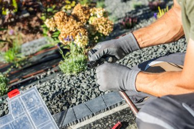 Bir bahçıvan sulama sisteminin bileşenlerini iyi korunmuş bir arka bahçede onarmaya odaklanır. Sıcak güneş ışığı arka plandaki canlı çiçekleri güçlendirir..