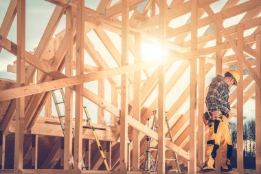 Bir inşaat işçisi parlak öğleden sonra güneşinin altında ahşap bir çerçeve kuruyor. Merdiven ve aletler etkin bölgenin etrafına saçılmış durumda..