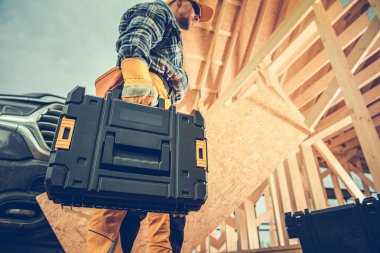 Kareli gömlekli ve kasklı bir inşaat işçisi şantiyede dururken alet çantası taşır. Tahta yapılar etrafını sarmış açık gökyüzünün ortasında..