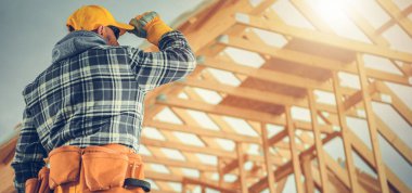 Ekose gömlek giyen bir inşaat işçisi ve yeni bir binanın iskeletini inceleyerek parlak güneş ışığına karşı şapkasını düzeltiyor..