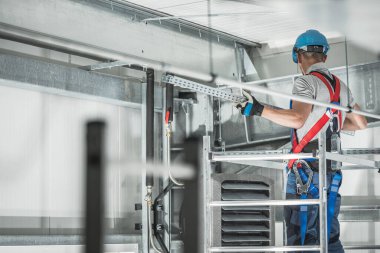 Bir inşaat işçisi, sanayi bölgesinde güvenlik önlemi alındığından emin olmak için sıkı bir şapka ve emniyet kemeri takarken ekipman kuruyor..