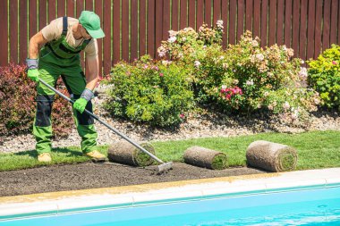 Bir bahçıvan, güneşli bir öğleden sonranın tadını çıkarırken bir yaz vahası için bahçeyi hazırlarken, bir yüzme havuzunun etrafına taze çimenler serer. Çiçeklerle çevrili..