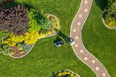 Bir insan bakımlı bir bahçede renkli çiçek yataklarıyla çevrili yemyeşil bir çim biçer. Sahne açık gökyüzü ile bir yaz gününü yakalıyor..