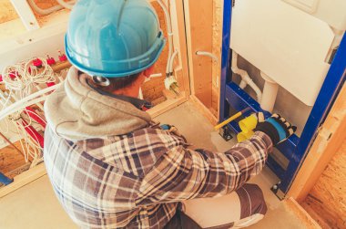 Mavi kask ve ekose ceket giyen bir inşaat işçisi, yeni inşa edilmiş tesisat tesisatına odaklanan boruları titizlikle birbirine bağlıyor..