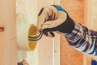 Eldiven ve ekose gömlek giyen yetenekli bir elektrikçi, renkli kabloları ahşap iskelet içindeki bir bağlantı kutusuna bağlar..