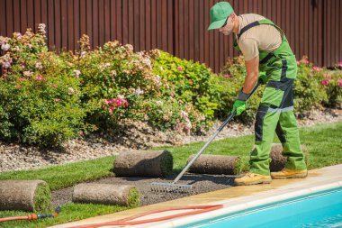 Güneşli bir günde bahçıvanın biri yüzme havuzunun yanına çimen seriyor. Renkli çiçekler arka plandaki canlı bahçe atmosferini güçlendirir.