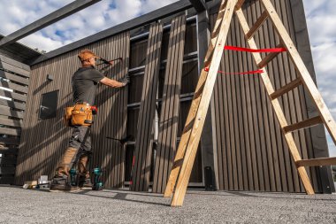 Yetenekli bir marangoz mavi bir gökyüzünün altındaki açık hava yapısına ahşap paneller yerleştirmeye çalışır. Dikkatli bir şekilde ölçüp hizalarken araçlar yakınlarda düzenlenir..