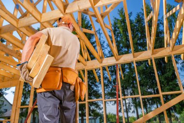 Bir işçi, açık mavi gökyüzü altında ahşap bir iskelet içinde büyük kereste parçaları taşıyor ve bir inşaat projesinin bir sonraki adımına hazırlanıyor..