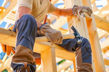 Bir marangoz, yapının üzerinde otururken tahta kirişleri korumak için çekiç kullanır. Parlak güneş ışığı arka plandaki inşaat alanını aydınlatıyor.