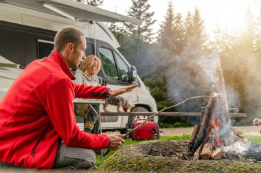Bir erkek ve bir kadın sıcak bir kamp ateşinde dinleniyor, yemek yapmaya hazırlanırken, akşam vakti uzun ağaçlarla çevrili sakin bir kamp yerinde..
