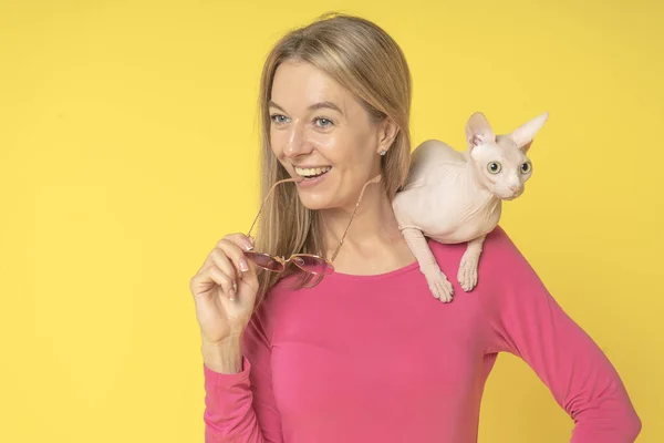 Happy and cheerful attractive woman with charming Sphynx Cat on shoulder looking in defferent sides. Joyful attractive woman with glasses in hand in pink dress isolated on yellow background.