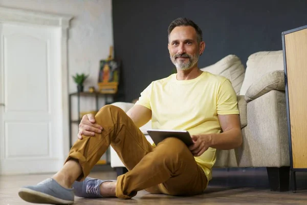 portrait of adult man dressed in yellow t-shirt sitting on the floor at home, leaning back on sofa with tablet pc in his hands. Looking at camera and smiling