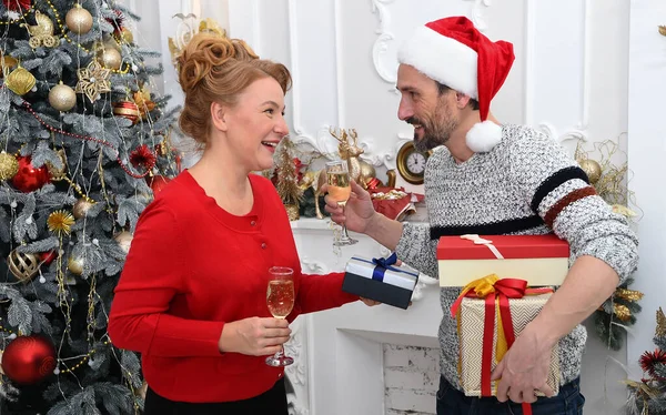 Gladsome couple celebrating New Year at home. Cheerful male tenderly looking at his pleased woman while holding present boxes and a glass of sparkling wine