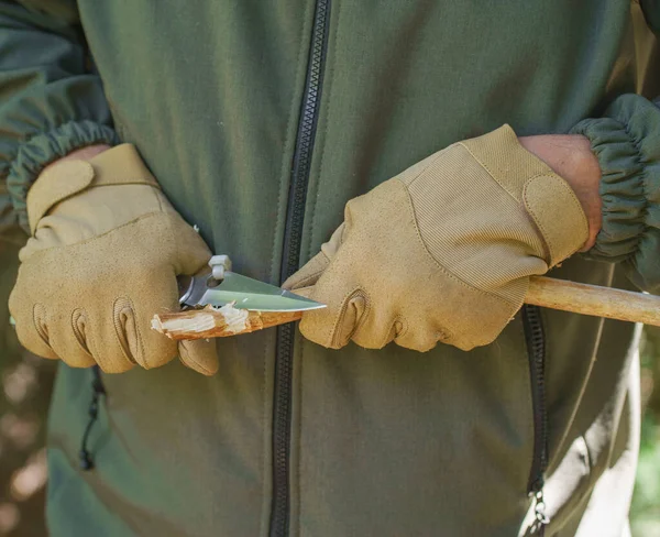 Close-up of knife in hands. hands in gloves cutting wooden branch outdoors with knife. Tourist man hands in tactical gloves with knife cut a wooden stick.