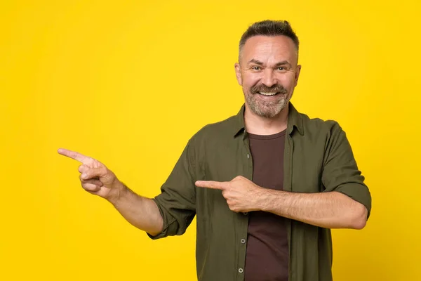 Senior grey haired stubble handsome man wearing green shirt standing isolated on yellow background smiling pointing with two hands and fingers to copy space