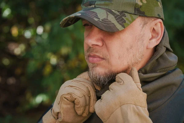 Close up of stubble man profile dressed in a military uniform camouflage cap and tactical gloves keeping jacket collar by two hands.