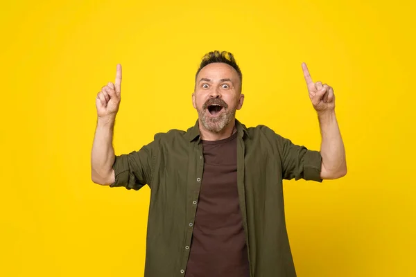Astonished with open mouth handsome mature bristle man with two hands pointing up to empty space above isolated yellow background dressed in green shirt and brown t-shirt