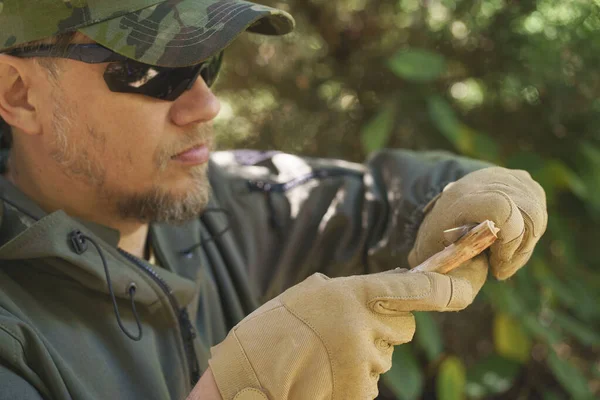 Close-up of man sharpening wood stick by knife in hands dressed in tactical gloves. Tourist man dressed in camouflage uniform, glasses and cap with knife in hands cut a wooden stick.