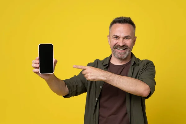 Attractive smiled bristle and mustached mature man 50s dressed in green shirt and brown t-shirt pointing to showing black screen of smartphone holded in hand on yellow background