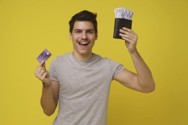 Mutlu 20 'li yaşlarda genç bir adam açık sarı arka planda izole edilmiş nakit para ve kredi kartı gösteriyor. Gençler için öğrenci kredisi. Debite banka kartından çekilen para