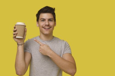 Genç, mutlu, yakışıklı, gri tişörtlü 25yo. Parmakla işaret ederek sarı arka planı kaplayan kahve fincanını gösteriyor.