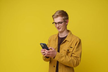 Sarı arka planda cep telefonu kullanan mutlu 20 'li yaşlarda genç bir adam. Akıllı telefon oyunu oynayan gülümseyen hippi, ecommerce mağazasından sarı ceketli bir cep telefonuyla online alışveriş yapan.
