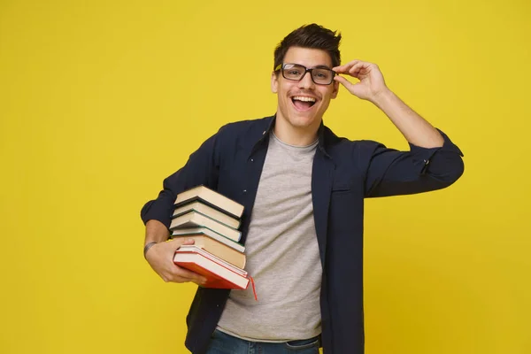 Happy smiling student standing with books in hand exciting holding his glasses dressed in blue shirt over yellow background. 25th young man holding books.