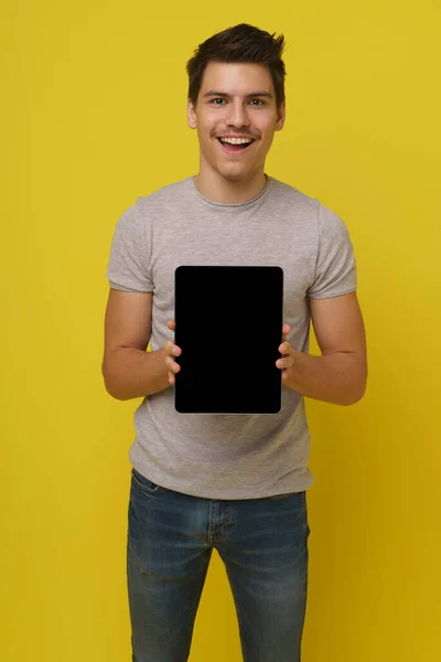 Gri tişörtlü mutlu 20 'li yaşlarda beyaz bir adam tablet bilgisayarını iki eliyle tutuyor ve ekranda siyah bir maket var. Sarı arka plan stüdyo resmine izole edilmiş modern teknoloji konsepti.