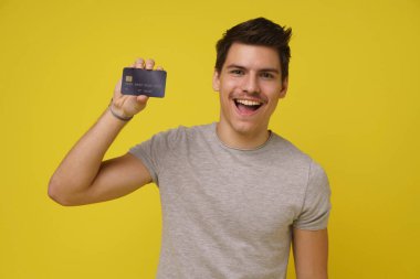 Çok heyecanlı, genç, esmer, 20 'li yaşlarda banka kartı olan, sarı arka planda izole edilmiş tişörtlü bir adamın portresi.