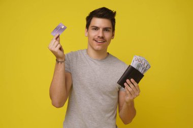Kredi kartı gösteren ve sarı arka planda izole edilmiş banknotlarla voleyi tutan mutlu genç adam.
