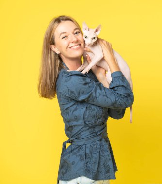 Evcil sfenks kedisi, sevimli genç bayana yanak yanağa sarılıyor. Sarışın, gülümseyen çift, arkadaşlık kadını ve kedi. Çekici kadın sarı arka planda soyutlanmış beyaz bir kediye sarılıyor.