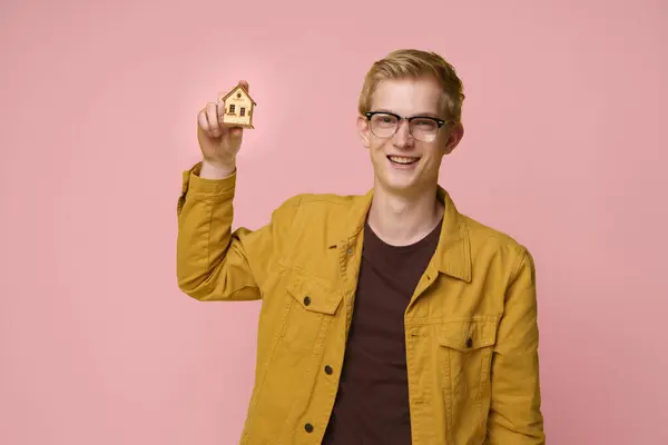 stock image A young man with glasses and a bright smile holds a miniature house in front of a pink background.