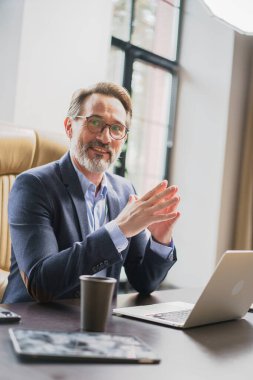 Modern bir ofis ortamında çalışırken masada dizüstü bilgisayarıyla kendini işine adamış olgun bir adamın portresi.