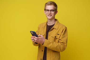 Gözlüklü ve sarı ceketli genç bir adam akıllı telefonunu açık sarı bir fon önünde kullanırken gülümsüyor..