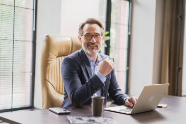 Başarılı ve kendinden emin bir işadamı bugün modern ofis ortamında özenle dizüstü bilgisayarı üzerinde çalışırken gülümsüyor.