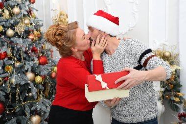 Şenlikli giyinmiş genç bir çift, güzel süslenmiş bir Noel ağacının yanında sevgi dolu bir öpücük ve hediye alışverişini paylaşıyor..