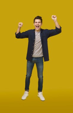 Kot pantolon ve düğmeli gömlek giyen genç bir adam saf sarı bir arka fona karşı duruyor, kolları zaferle havada, yüzünde geniş bir sırıtış.