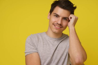Gri tişörtlü genç bir adam, parlak sarı bir arka plana karşı düşünceli bir ifadeyle doğrudan kameraya bakıyor..