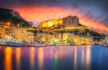 Bonifacio, Korsika 'nın manzarası. Parlak bir günbatımı gökyüzü, sakin bir limanda yatlar ve geceleri aydınlatılmış tarihi bir kale..